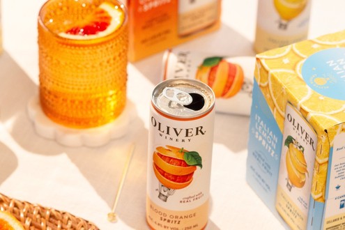 Open Oliver Winery Blood Orange Spritz can with a glass of spritz and an Italian Lemon Spritz 4-pack on a sunny table