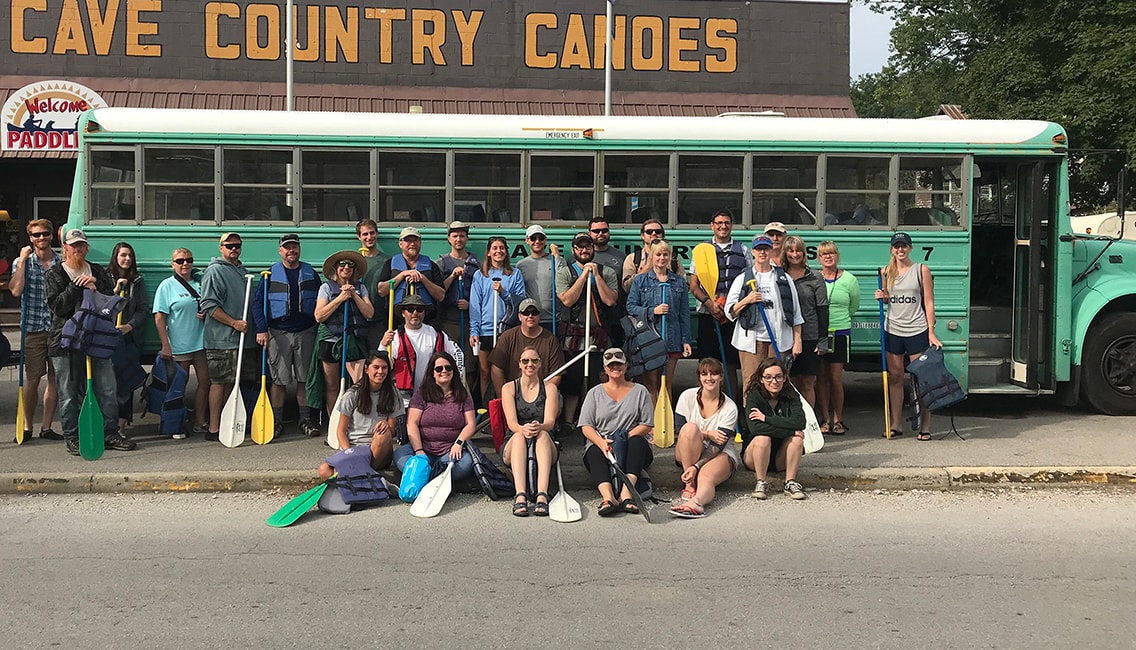 Large group of Oliver Winery staff members gathered with oars and life jackets in front of a bus and a “cave country canoes” sign.