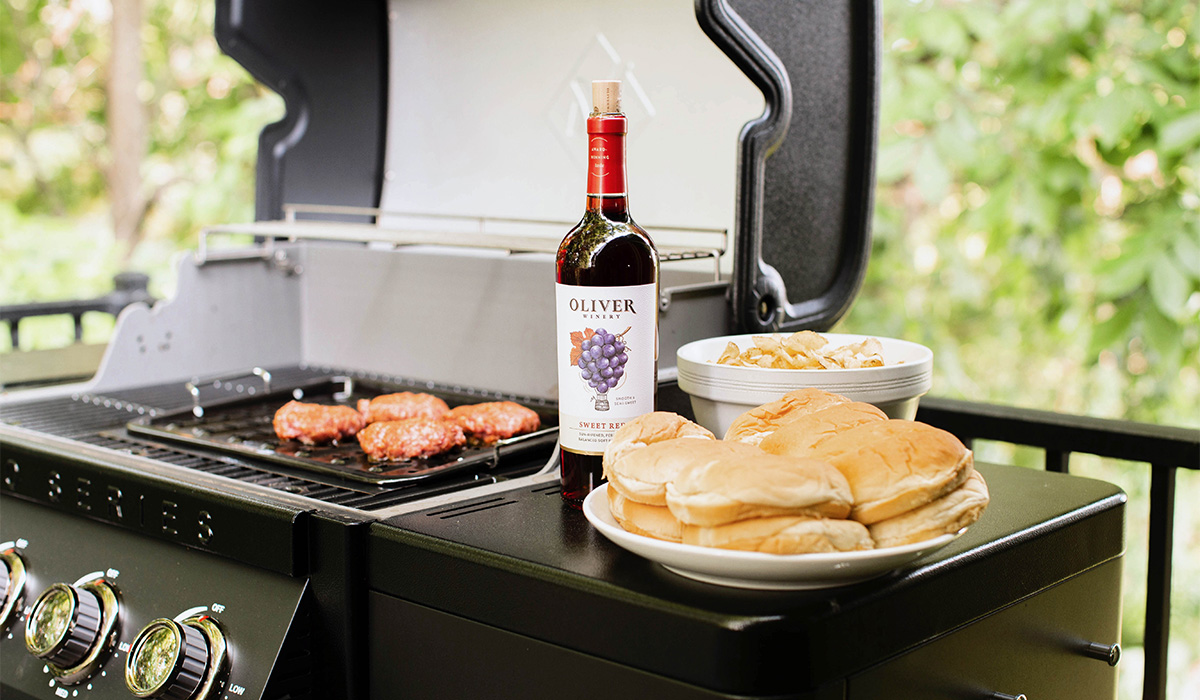 Bottle of Oliver Sweet Red on a table with steak on the patio by a pool.