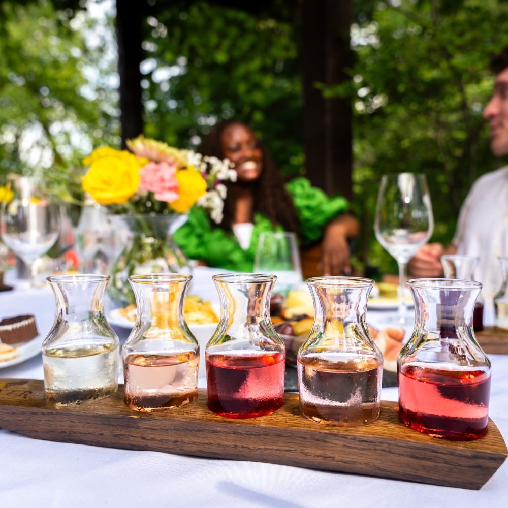 A wine flight of five small Oliver Winery carafes on a wooden board at an outdoor table, with guests dining behind it.