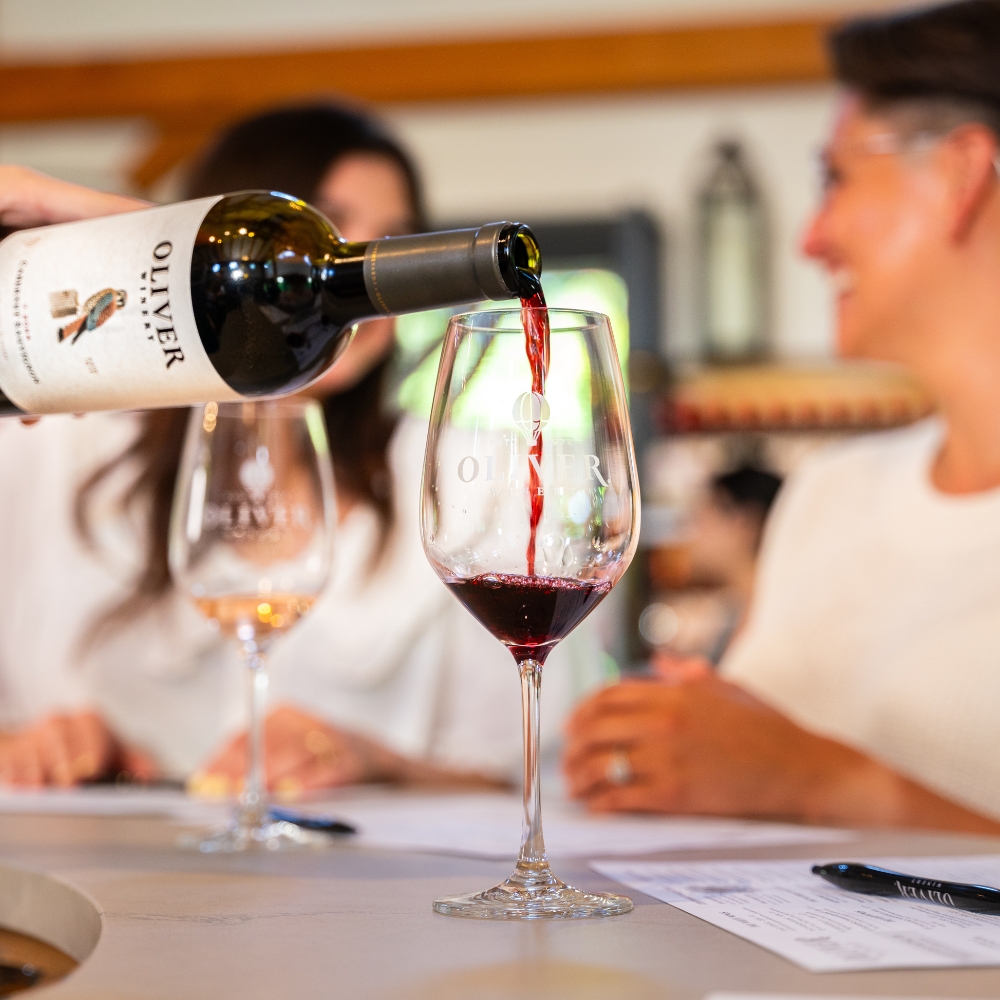 Red wine being poured into an Oliver Winery logo glass at the tasting bar as two guests look on.