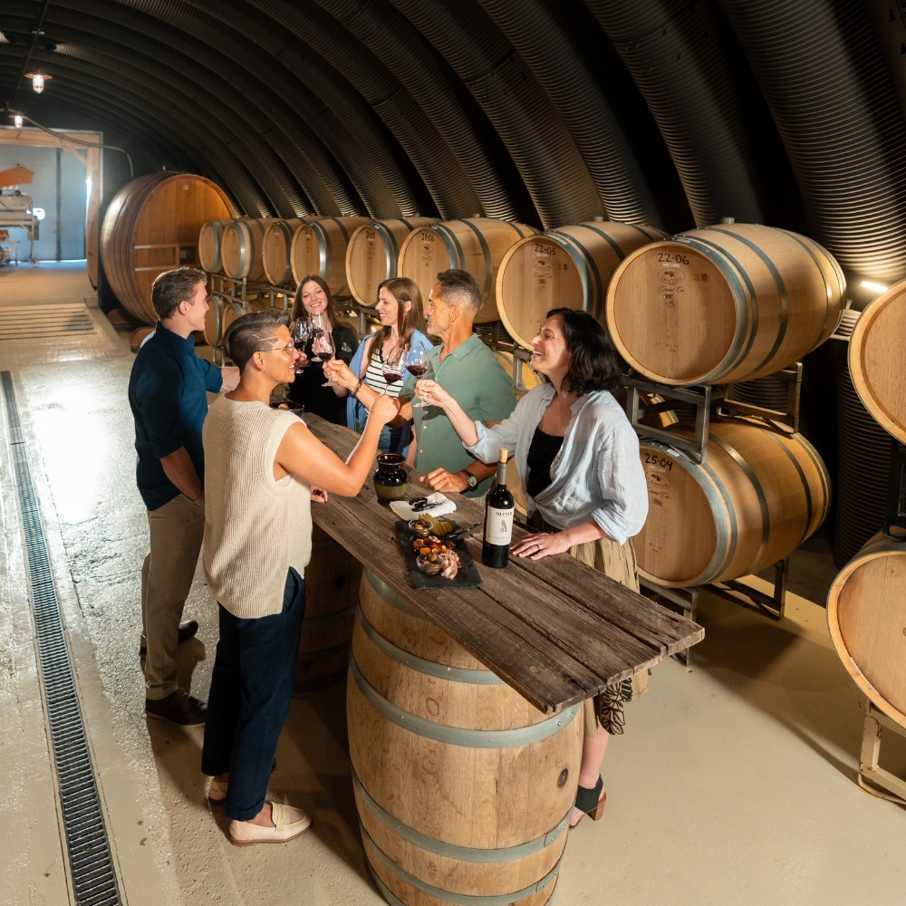 A tour group tastes red wine around a barrel-top table in the Oliver Winery barrel cellar.