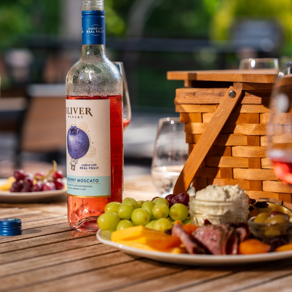 A chilled bottle of Oliver Blueberry Moscato on an outdoor table beside a charcuterie plate and picnic basket.