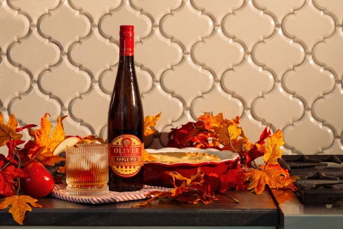 Bottle of Apple Pie wine from Oliver Winery on a countertop surrounded by autumn leaves, with a poured glass and a fresh pie.