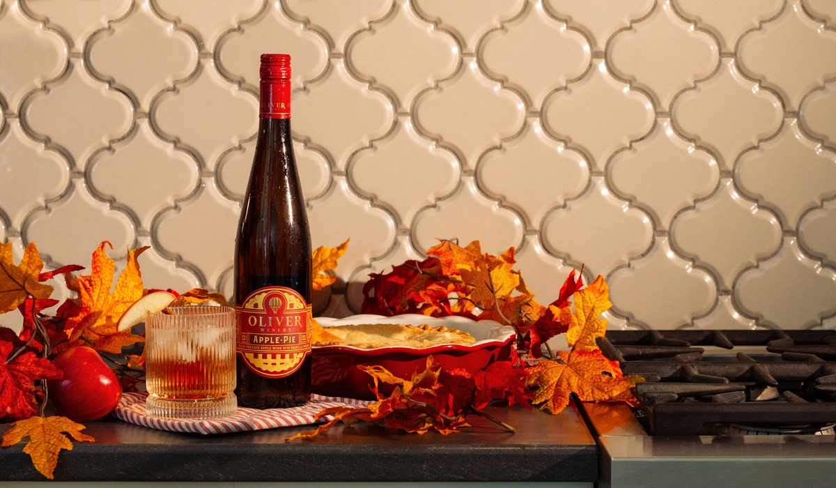 Bottle of Apple Pie wine from Oliver Winery on a countertop surrounded by autumn leaves, with a poured glass and a fresh pie.