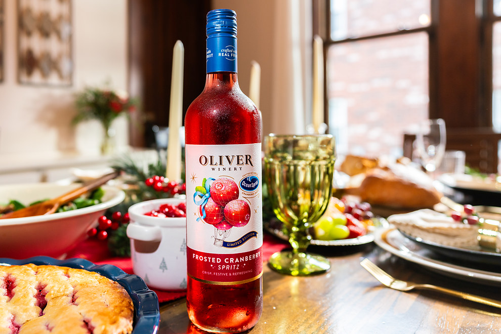 A bottle of Oliver Winery Frosted Cranberry Spritz on a holiday table with a cranberry pie, a bowl of cranberry sauce, a salad, taper candles and a green goblet.