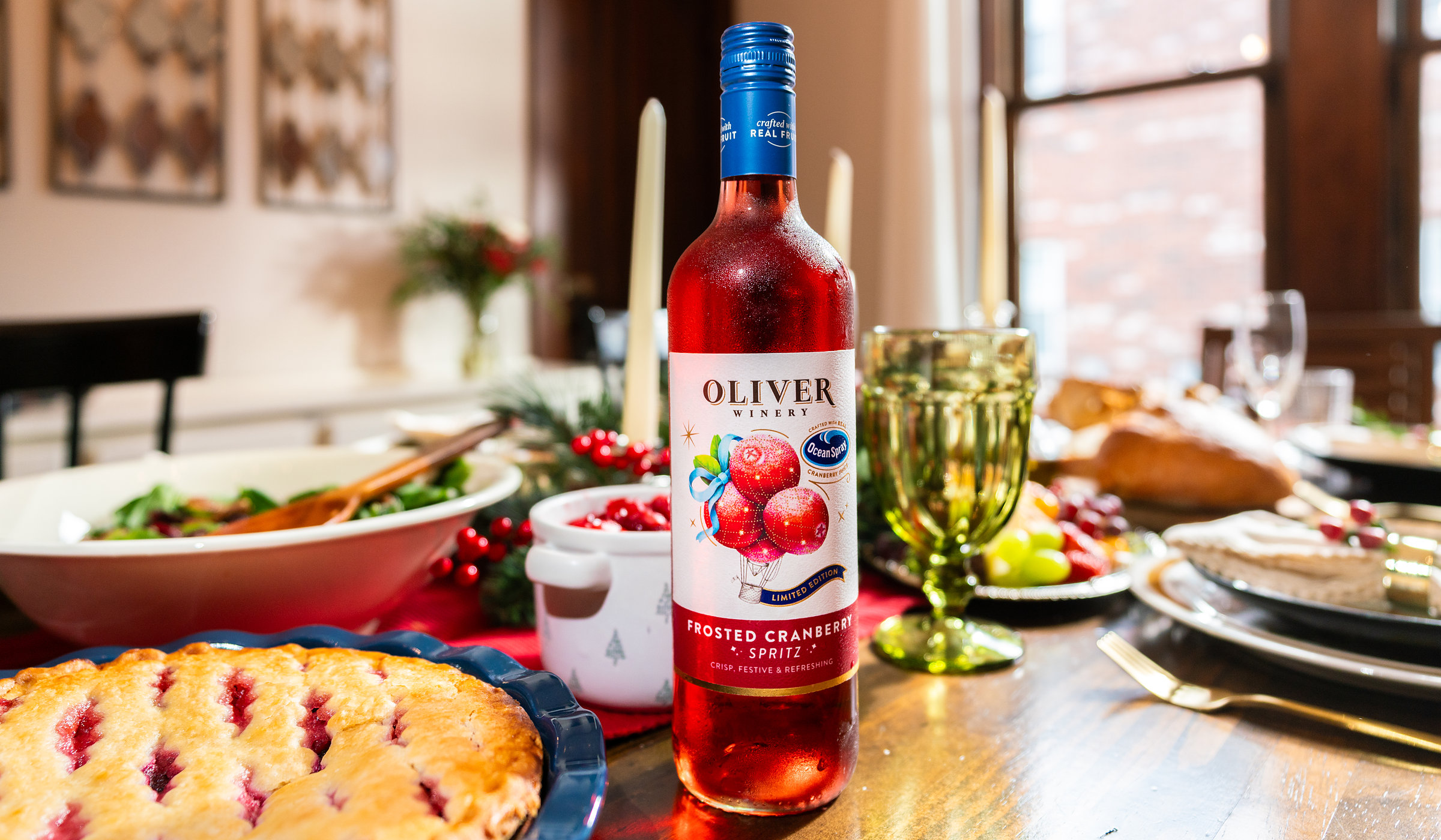A bottle of Oliver Winery Frosted Cranberry Spritz on a holiday table with a cranberry pie, a bowl of cranberry sauce, a salad, taper candles and a green goblet.