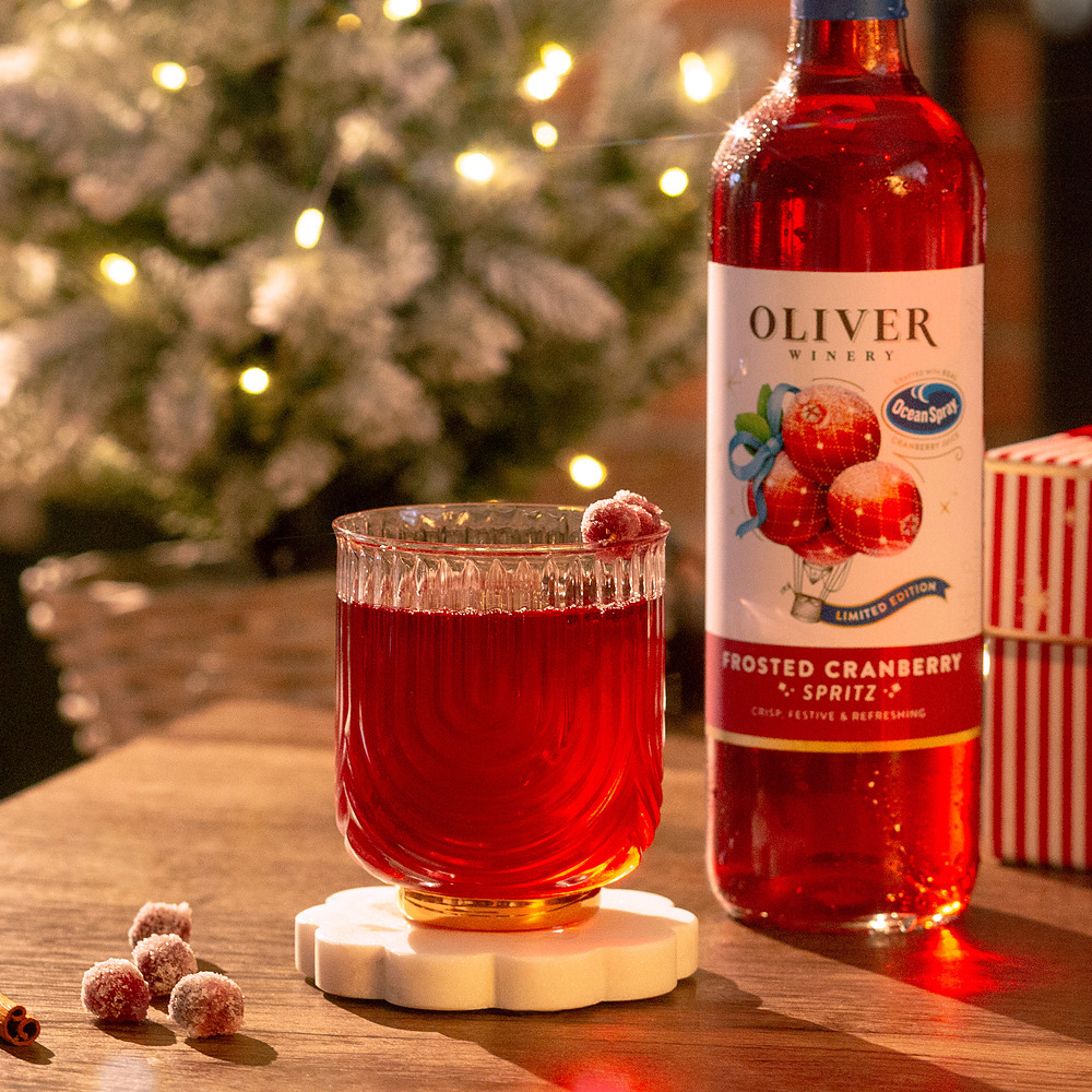 A glass of Oliver Winery Frosted Cranberry Spritz garnished with frosted cranberries, beside the bottle, a striped gift box and a lit Christmas tree.
