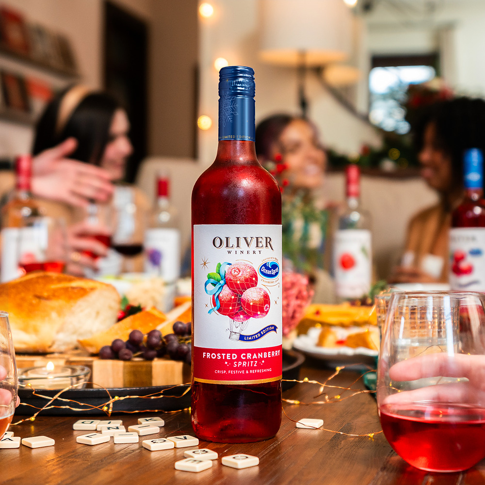 A bottle of Oliver Winery Frosted Cranberry Spritz on a holiday table with fairy lights, bread and a charcuterie board, with friends gathered behind it.