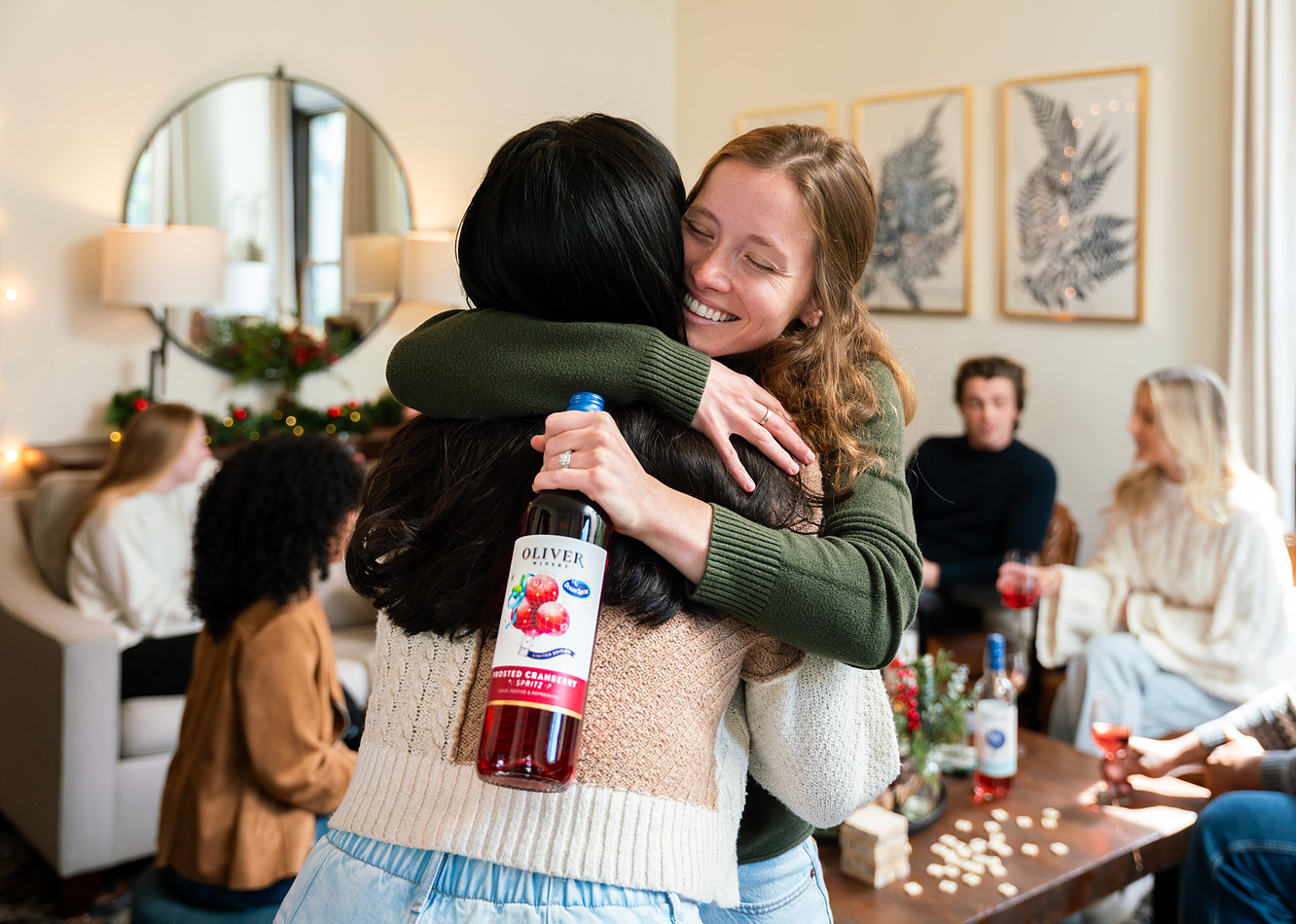 Two friends hugging at a holiday party, one holding a bottle of Oliver Winery Frosted Cranberry Spritz, with other guests talking on couches behind them.