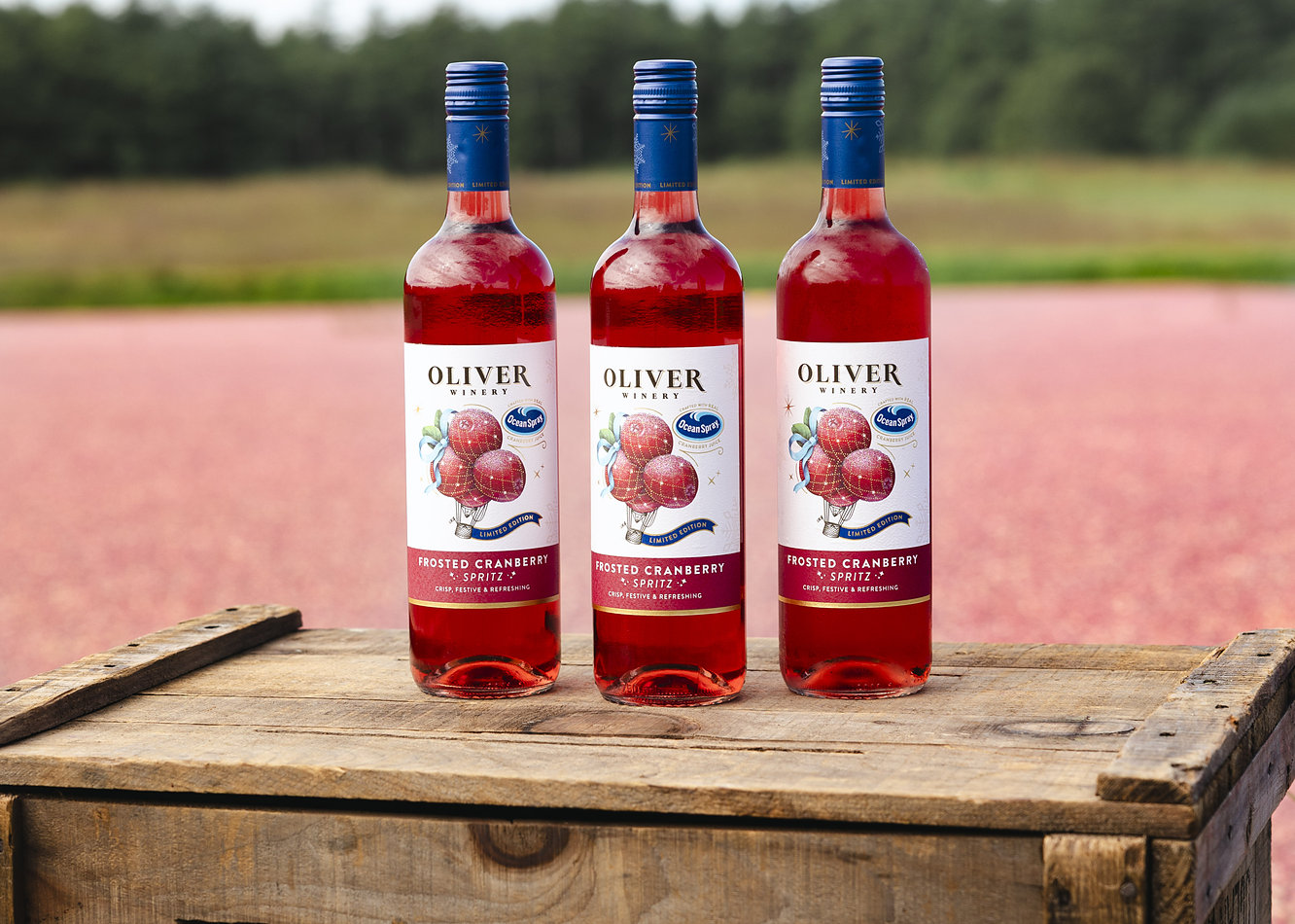 Three bottles of Oliver Winery Frosted Cranberry Spritz on a wooden crate beside a flooded cranberry bog at harvest.