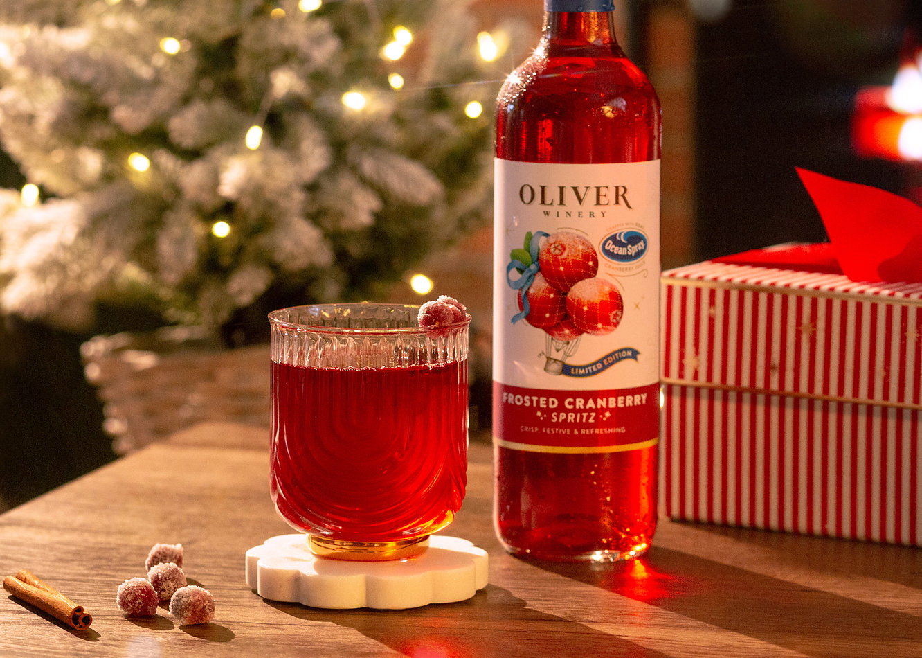 A glass of Oliver Winery Frosted Cranberry Spritz garnished with frosted cranberries, beside the bottle, a wrapped gift and a lit Christmas tree.