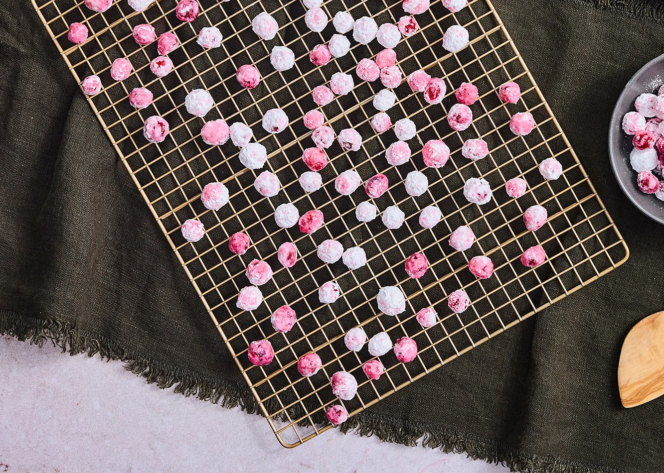Frosted cranberries drying on a gold cooling rack over a dark green linen cloth, with a bowl of finished cranberries and a wooden spoon beside it.