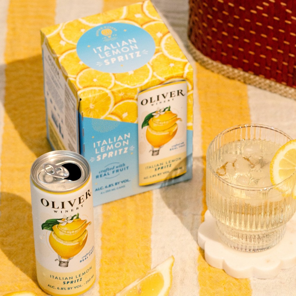 Oliver Winery Italian Lemon Spritz can and 4-pack carton on a yellow striped blanket, with a glass of spritz over ice and lemon slices.