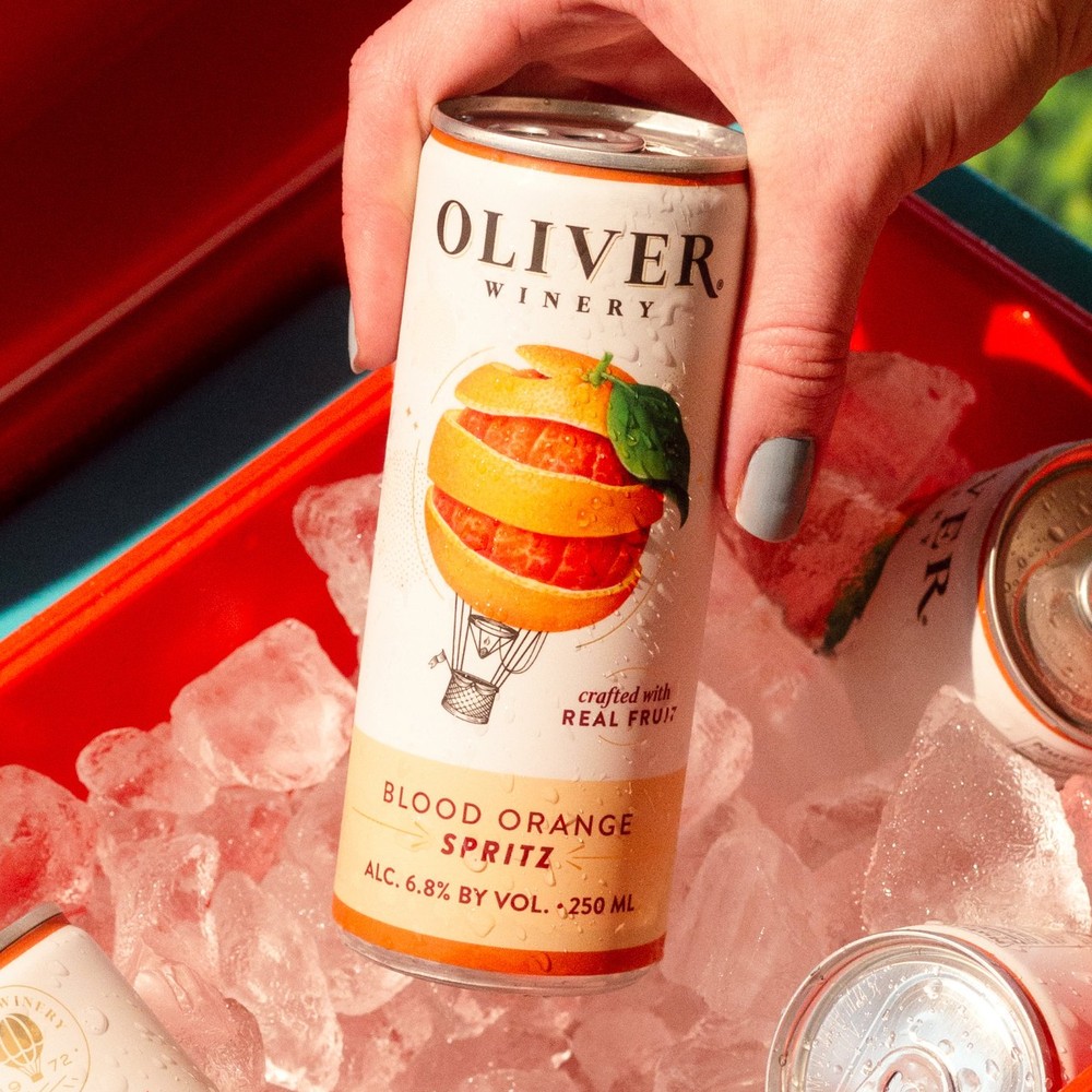 A hand lifts an Oliver Winery Blood Orange Spritz can out of a cooler full of ice, with more cans nestled around it.