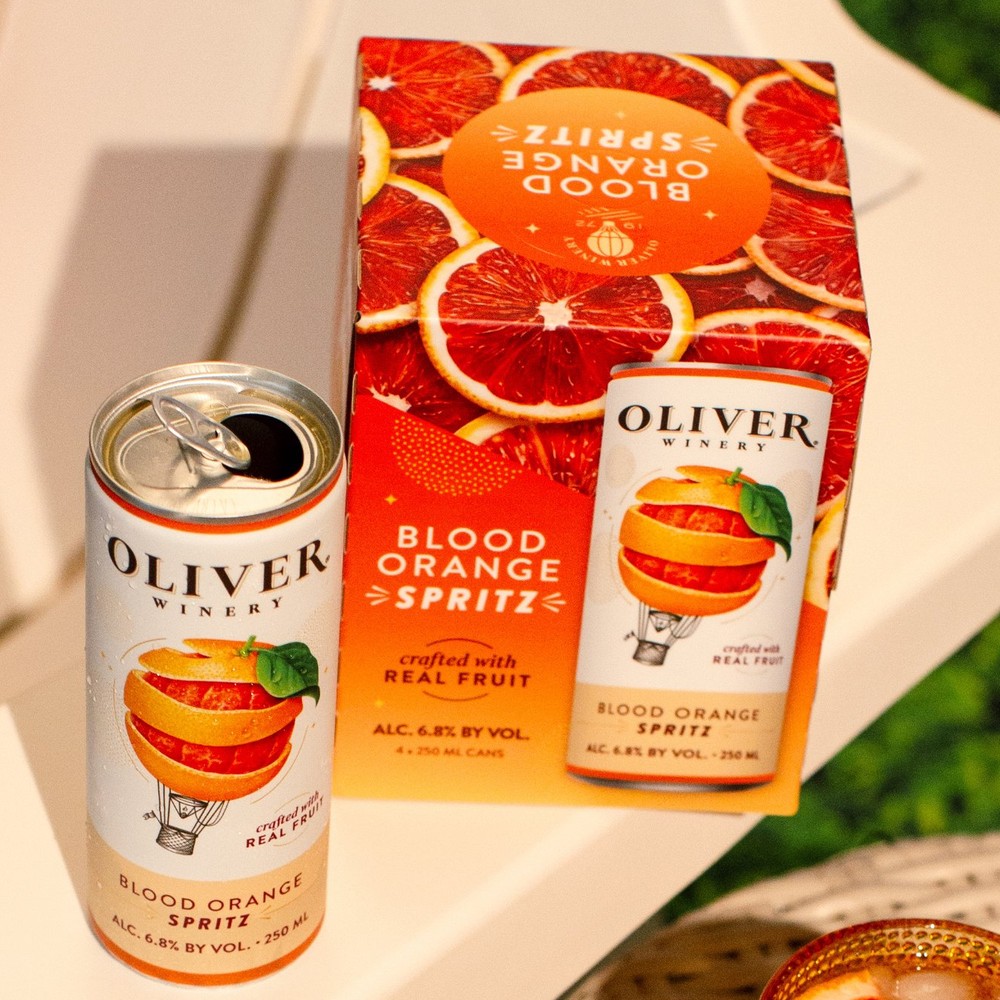 An open Oliver Winery Blood Orange Spritz can next to its 4-pack carton on a white patio chair, with a glass of spritz nearby.