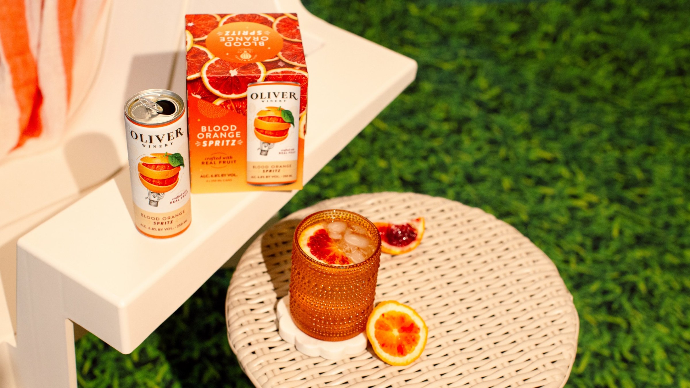 Oliver Winery Blood Orange Spritz can and 4-pack on a white patio chair, with a glass of spritz over ice on a wicker table