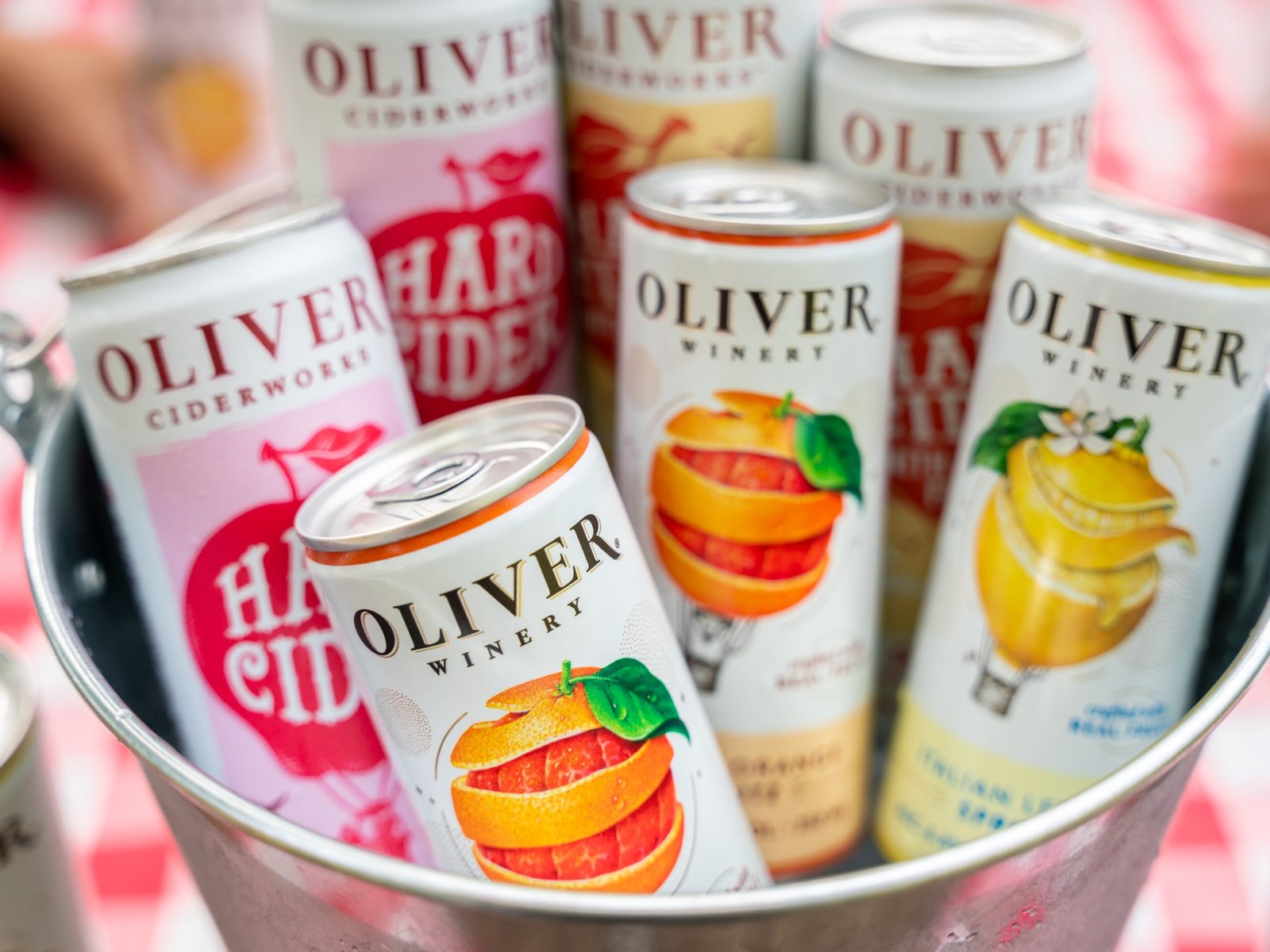 Oliver Winery Blood Orange and Italian Lemon Spritz cans with Oliver Ciderworks Hard Cider in a metal bucket on a red gingham table