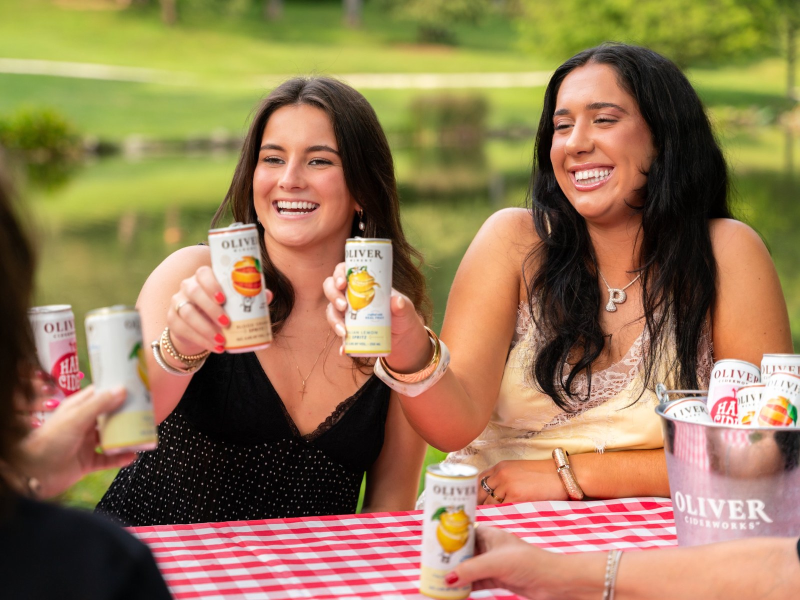Friends raising Oliver Blood Orange and Italian Lemon Spritz cans in a toast at a lakeside picnic table with a red gingham cloth